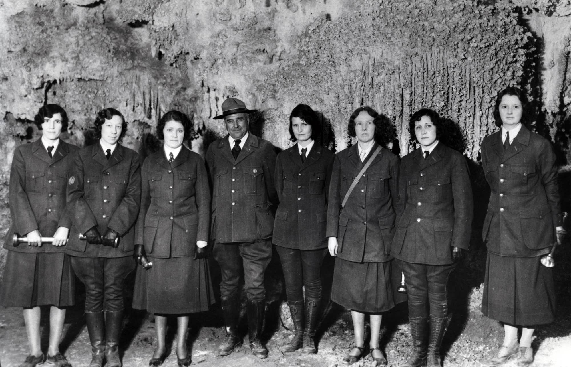 Seven women pose with Thomas Boles inside a cave. They all wear NPS uniforms. Only Boles wears a broad brim hat and a round superintendent’s badge.